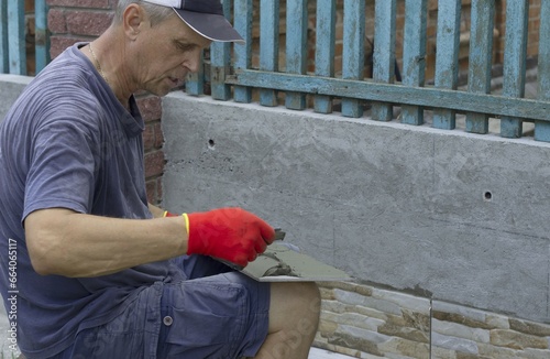 A male construction worker applies glue to the facing tile to lay it on the enclosing wall of the building from the outside.