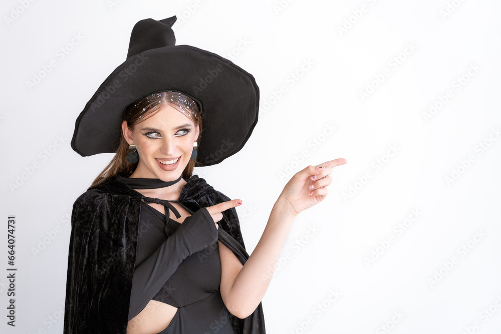 Smiling beautiful woman in black hat and cloak points her index finger to the side. White background. Space for text. Black Friday. Halloween party Smiling beautiful woman in black hat and cloak points her index finger to the side. White background. Space for text. Black Friday. Halloween party