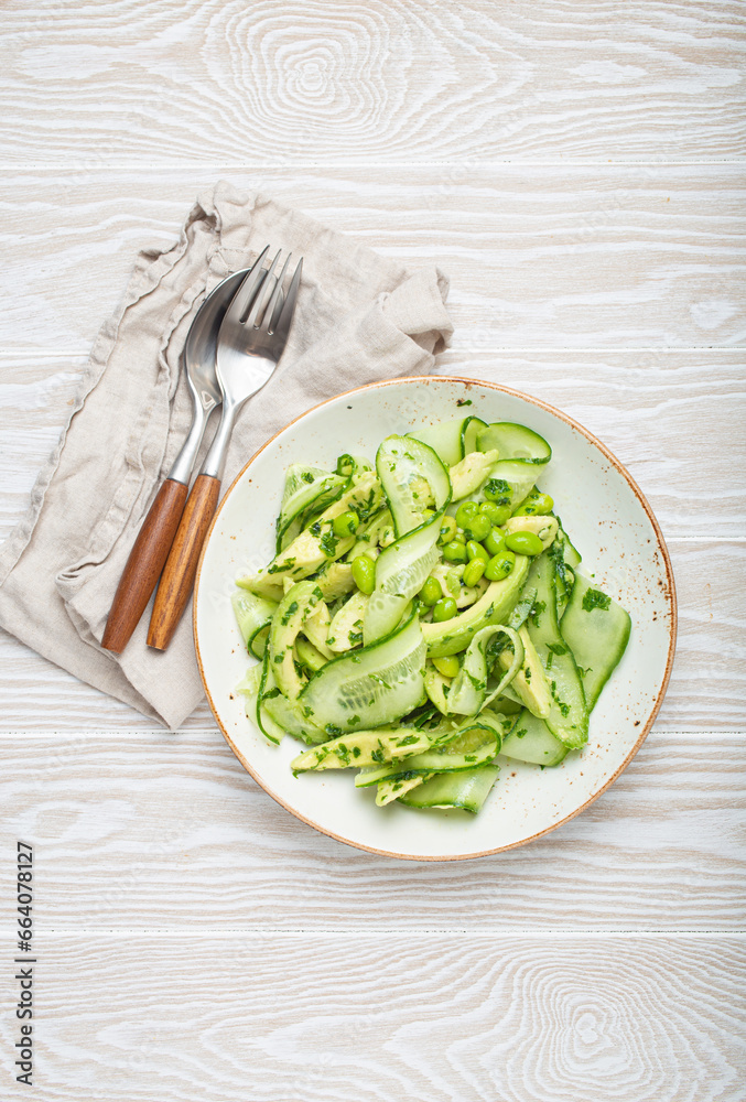 Healthy vegan green avocado salad bowl with sliced cucumbers, edamame beans, olive oil and herbs on ceramic plate top view on white wooden rustic table background Healthy vegan green avocado salad bowl with sliced cucumbers, edamame beans, olive oil and herbs on ceramic plate top view on white wooden rustic table background