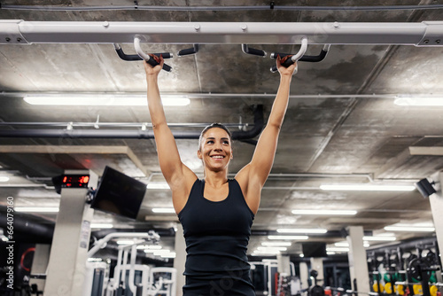 Portrait of a happy sportswoman doing pullups in a gym.