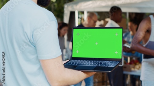 Caucasian man shown in a close-up holding digital laptop with isolated chromakey template. Male volunteer at an outdoor food bank grasping wireless computer displaying blank green screen.