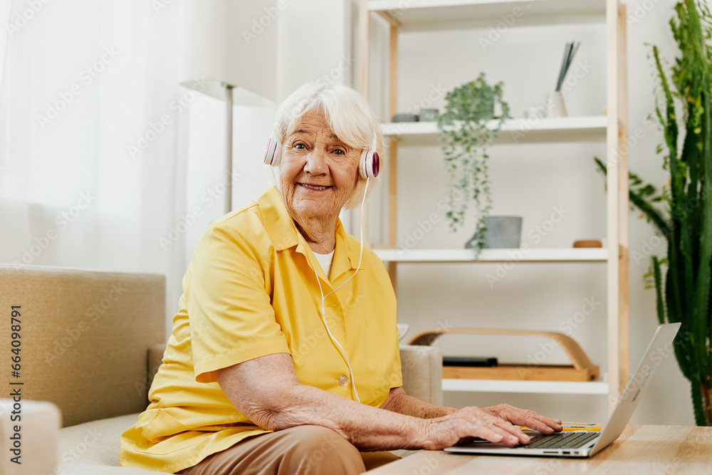 An elderly woman with headphones and a laptop sits on the couch at home and works. An elderly woman with headphones and a laptop sits on the couch at home and works.