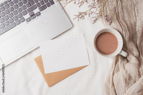 Coffee cup, laptop computer, envelope with blank card and small flowers on white linen. Cozy workplace, breakfast, comfort, hygge concept. Top view, flat lay, mockup