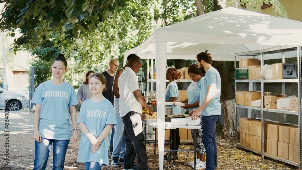 Caucasian mother and her daughter taking part in a hunger relief initiative at a food bank. Multiethnic volunteers in blue t-shirts outdoors ready to help the need and homeless people. Slow-mo. Caucasian mother and her daughter taking part in a hunger relief initiative at a food bank. Multiethnic volunteers in blue t-shirts outdoors ready to help the need and homeless people. Slow-mo.