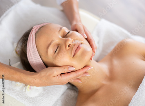 Female client is resting during massage face skin. Female physiotherapist performs restorative stroking during spa care.