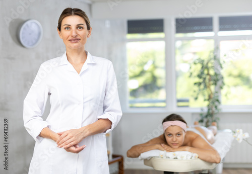 Positive confident young professional masseuse standing in cozy massage room with female client lying on bed and waiting for procedure