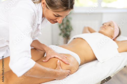 Female masseur professional performs stroking and patting during anti-cellulite spa procedure of front thigh area and leg for girl client. Therapeutic restorative massage.