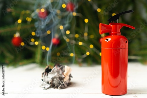 The fire extinguisher stands on a white table, against the background of a Christmas tree and smoke, decorated with colorful electric garlands, at home. The concept of fire safety