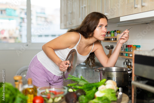 Young woman making and tasting healthy vegetable soup in kitchen