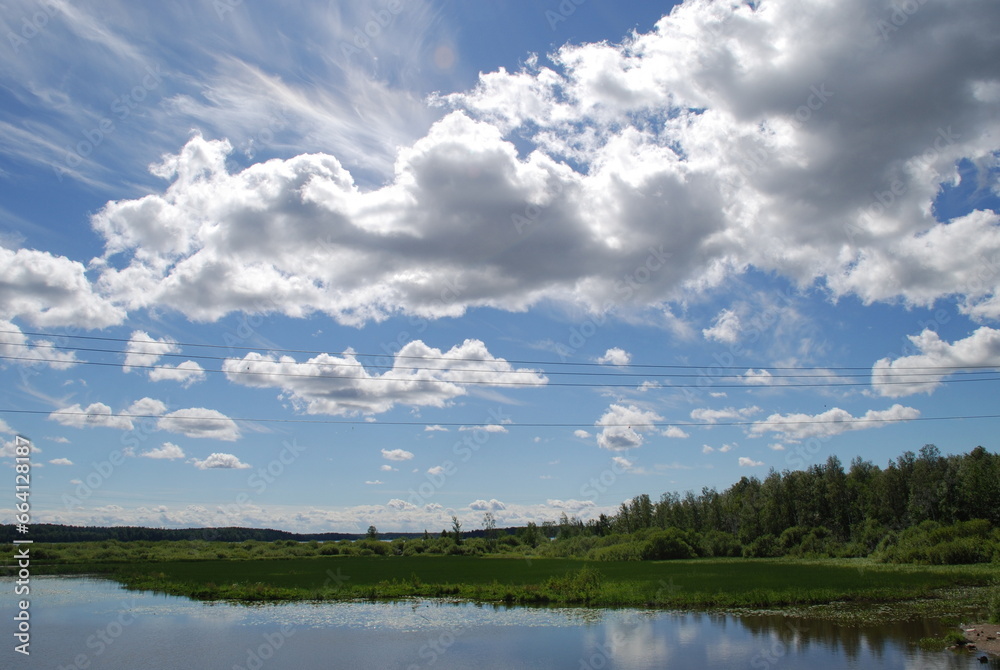 Lake under a cloudy sky. Forest lake with small waves on the surface, under a light blue sky with white cumulus and cirrus clouds. A forest with green foliage grows along the shores of the lake. Lake under a cloudy sky. Forest lake with small waves on the surface, under a light blue sky with white cumulus and cirrus clouds. A forest with green foliage grows along the shores of the lake.