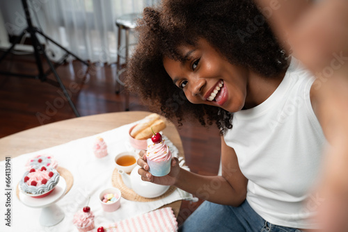 Beautiful young African blogger presenting piece of cupcake in concept special cuisine with selfie on smartphone. Content creating of social media with favorite sweets bakery dish. Tastemaker.