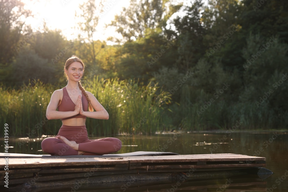 Fototapeta premium Beautiful happy woman practicing Padmasana on yoga mat on wooden pier near pond, space for text. Lotus pose