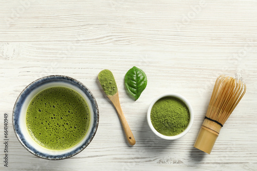Cup of fresh matcha tea, green powder and bamboo whisk on white wooden table, flat lay. Space for text