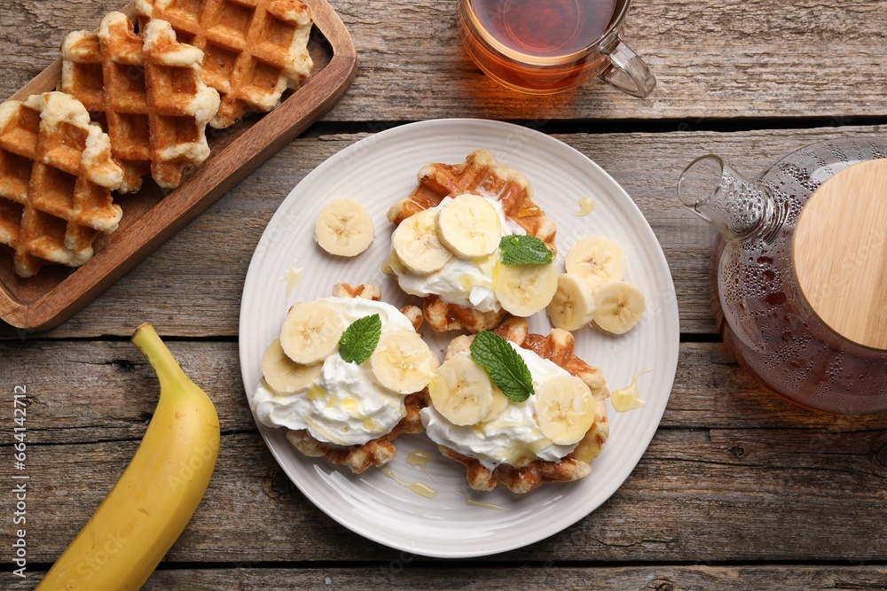 Delicious Belgian waffles with banana and whipped cream served on wooden table, flat lay