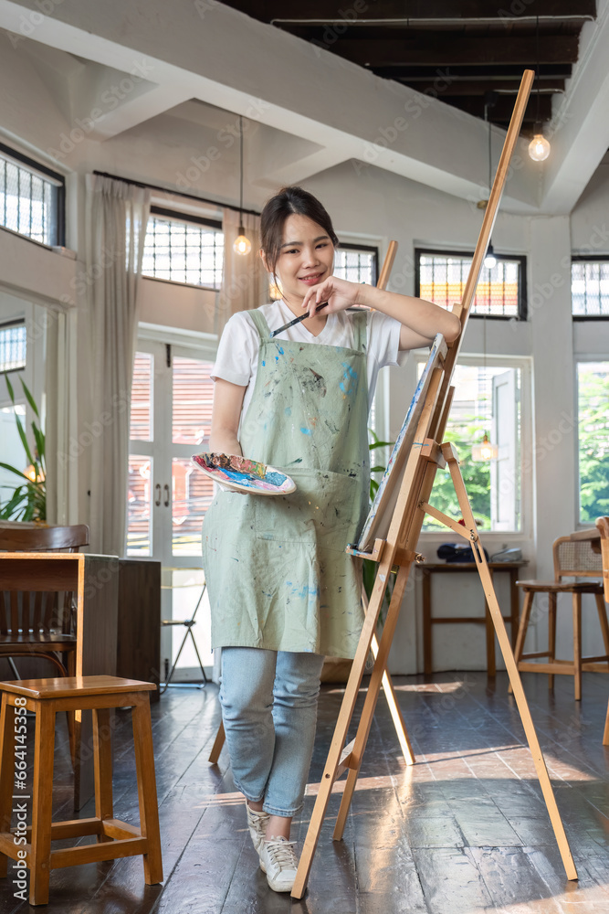 Portrait of young woman painting holding a palette and paintbrush in front of a canvas in a painting studio.