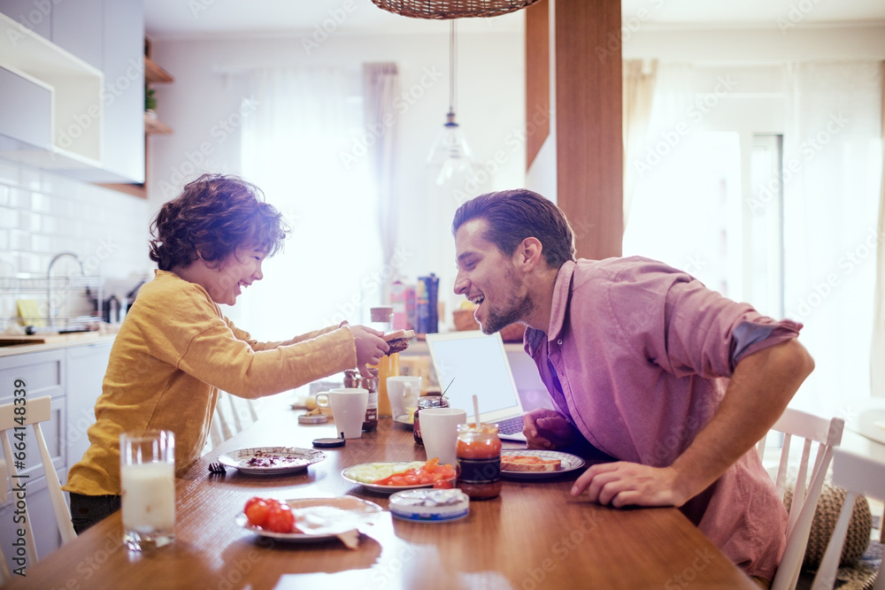 Happy young boy feeding his dad at the breakfast table in the kitchen