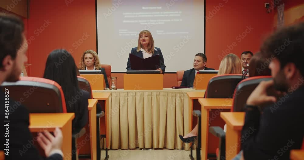 A group of students and professors gather for a graduation exam at the faculty. The atmosphere is tense as a student defends her diploma, showcasing her knowledge. In the audience, two students engage A group of students and professors gather for a graduation exam at the faculty. The atmosphere is tense as a student defends her diploma, showcasing her knowledge. In the audience, two students engage