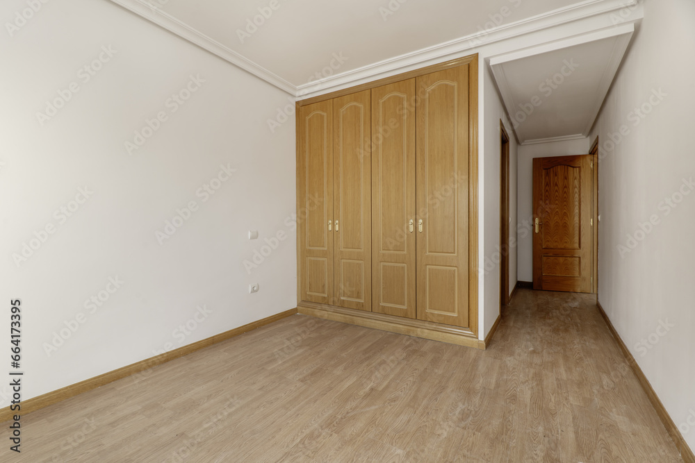 Empty bedroom with three-door built-in oak wardrobe with handles, skirting board of the same material, en-suite bathroom with wooden doors and plaster moldings on the ceiling Empty bedroom with three-door built-in oak wardrobe with handles, skirting board of the same material, en-suite bathroom with wooden doors and plaster moldings on the ceiling