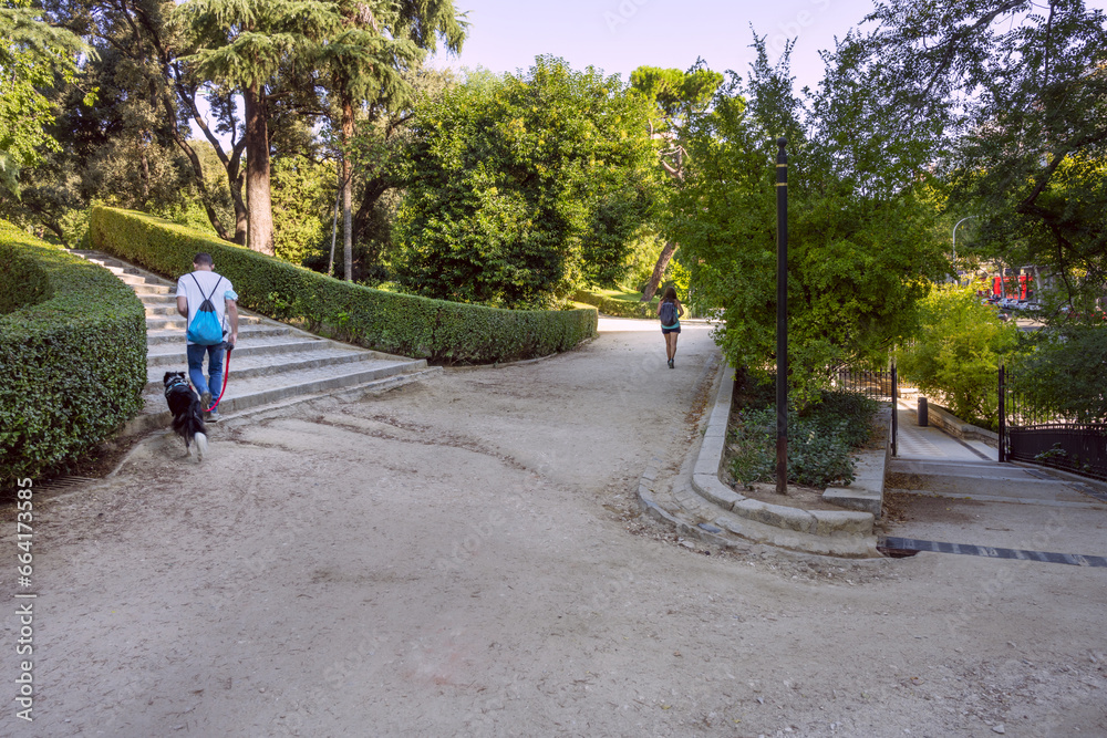 an urban park with dirt paths, people and animals walking, stone stairs, metal fence and many trees