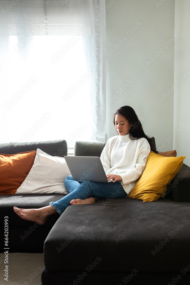 Young Asian woman working with laptop at home sitting on the couch in cozy apartment living room. Copy space. Young Asian woman working with laptop at home sitting on the couch in cozy apartment living room. Copy space.