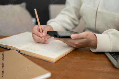 Close-up image of a woman using her smartphone and taking notes in her book at her desk.