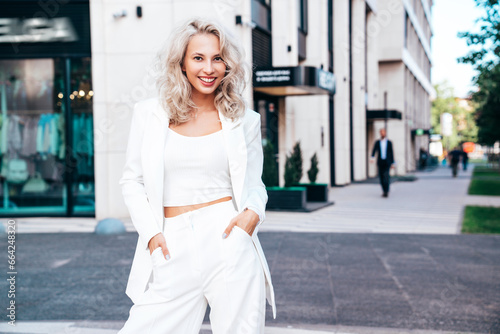 Young beautiful blond woman wearing nice trendy white suit jacket. Smiling model posing in the street at sunny day. Fashionable female outdoors. Cheerful and happy