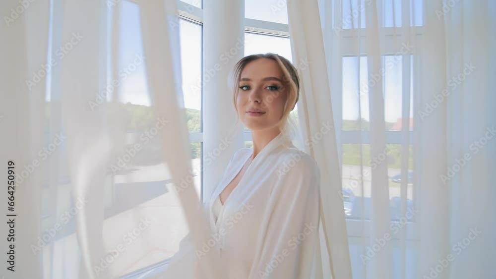 A happy bride in a white robe stands by the window behind the curtain, turns around and covers herself with the curtain. Wedding morning and preparation of the bride for the wedding day. A happy bride in a white robe stands by the window behind the curtain, turns around and covers herself with the curtain. Wedding morning and preparation of the bride for the wedding day.