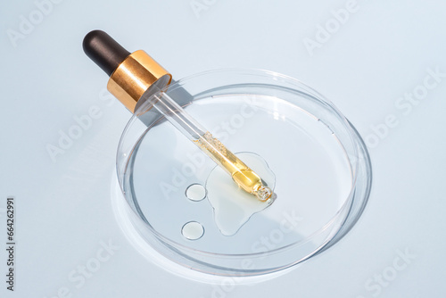 Yellow beauty face oil dripping from gold dropper on blue background in petri dish. Skin care cosmetic advertisement with copy space