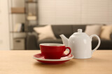 Red cup of fresh tea and teapot on wooden table in room