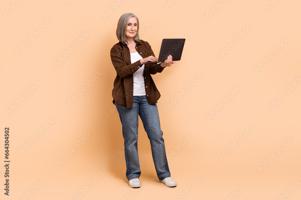 Full size body cadre of confident programmer old woman coding devops use netbook wear jeans brown shirt isolated on beige color background Full size body cadre of confident programmer old woman coding devops use netbook wear jeans brown shirt isolated on beige color background