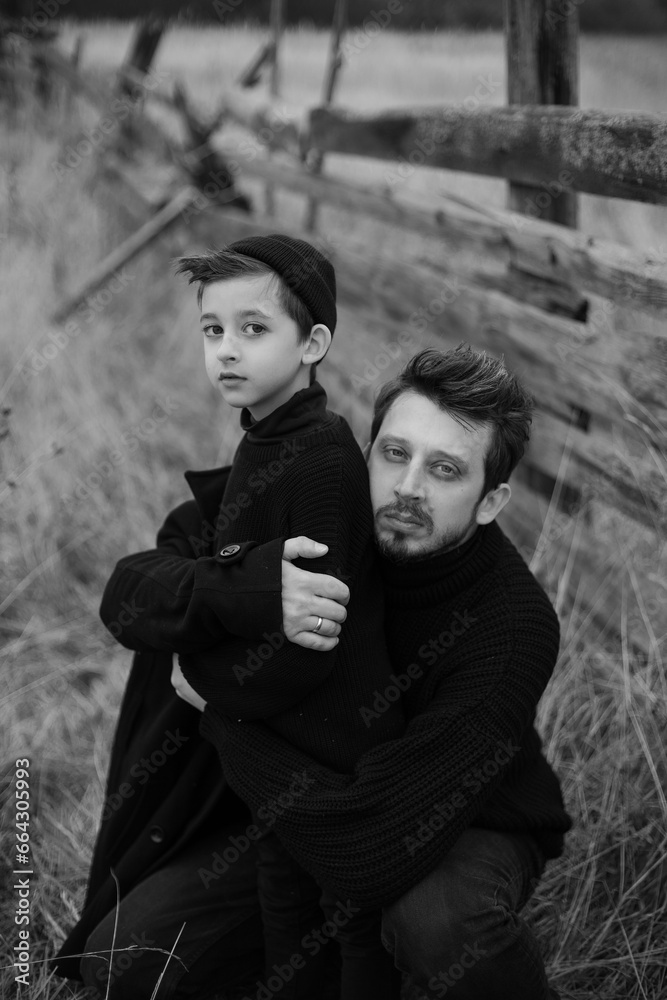 portrait stylish father and son in black clothes stand in a field with an old wooden horse fence in the fall. black and white