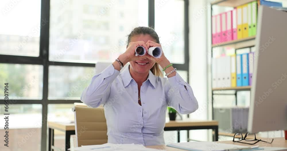 Playful businesswoman sits at table and looks at roll of paper through binoculars. Observing colleagues in the office and searching for business ideas