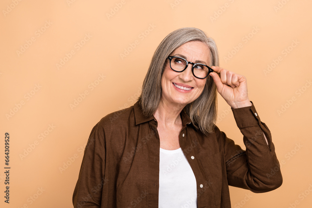 Photo of toothy beaming woman with gray hairdo dressed brown shirt touching glasses look empty space isolated on pastel color background