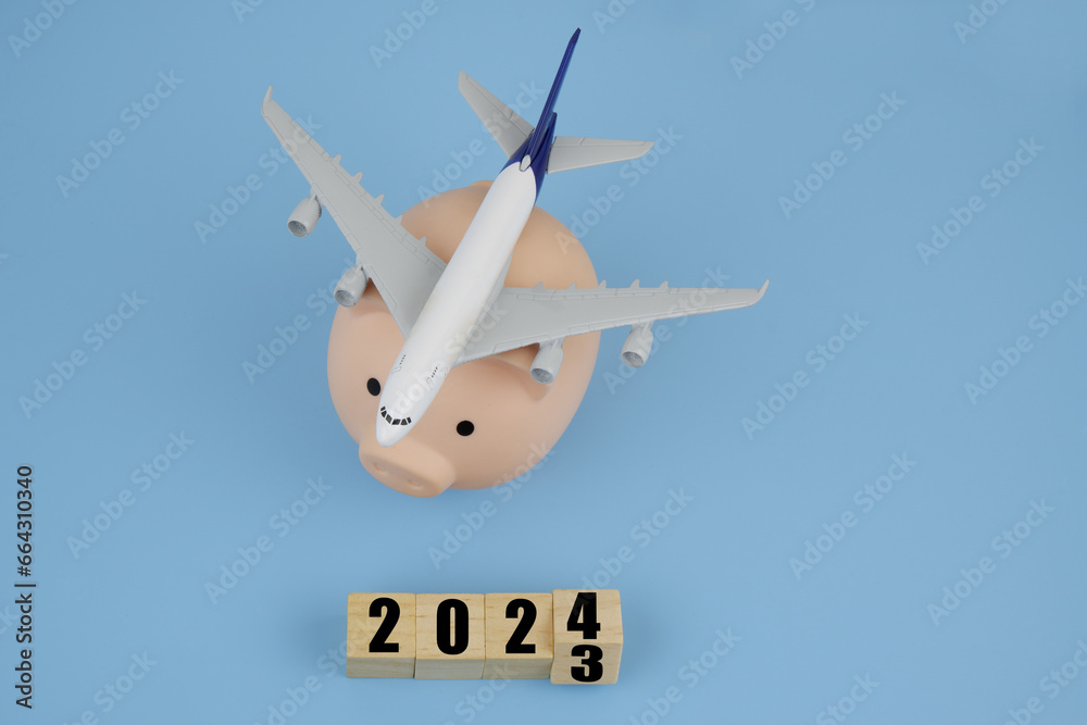 Airplane on piggy bank and wooden cubes with 2023 and 2024 on blue background. Airplane on piggy bank and wooden cubes with 2023 and 2024 on blue background.