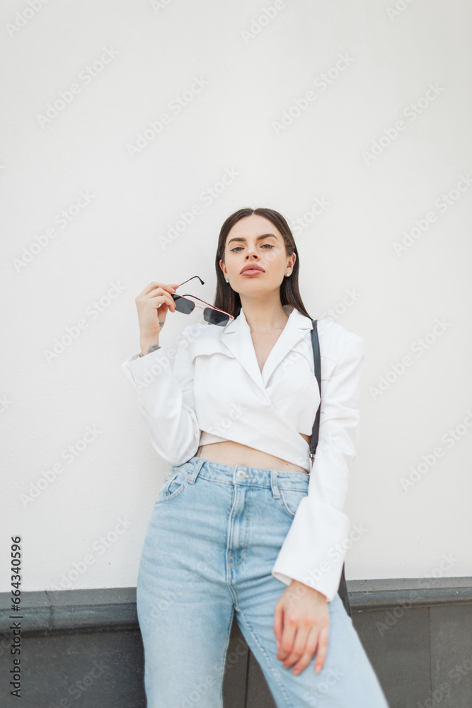 Pretty beautiful young woman with sunglasses in trendy fashion outfit with white blazer and vintage blue jeans stands near a white building on the street Pretty beautiful young woman with sunglasses in trendy fashion outfit with white blazer and vintage blue jeans stands near a white building on the street
