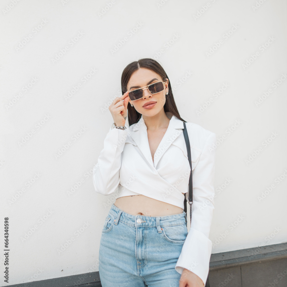 Beautiful stylish fresh urban woman in fashion clothes with a white jacket and vintage blue jeans puts on sunglasses near a white wall in the city Beautiful stylish fresh urban woman in fashion clothes with a white jacket and vintage blue jeans puts on sunglasses near a white wall in the city