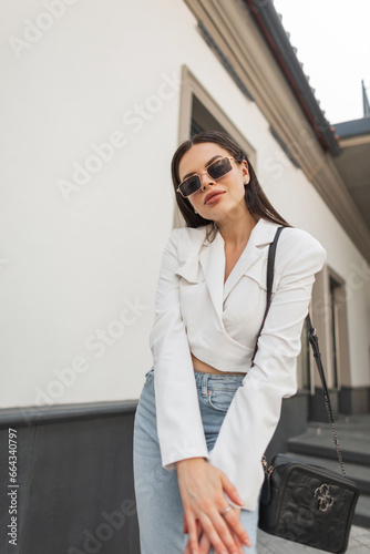 Trendy stylish beautiful young woman model with sunglasses in fashion outfit with a fashionable bag poses in the city