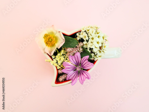 White heart shaped coffee cup with colorful spring flowers against pastel pink background. Natural minimal concept. Creative spring idea. Flat lay. Valentine's day or Woman's day desing.