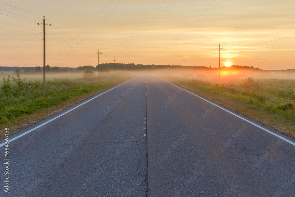 A distant suburban highway on a foggy summer morning. Beautiful foggy landscape with the sun rising from the horizon.
