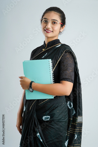 Portrait of Indian lady teacher in saree stands against white or blackboard , conducting online class using Camera, internet and lights