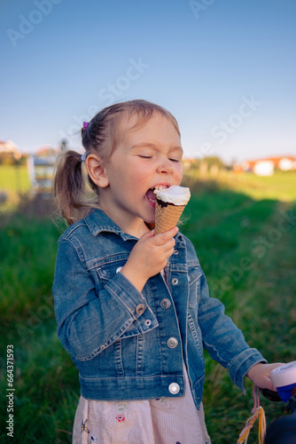 Little beautiful girl of European appearance eating ice cream while sitting on her pink bicycle. Girl with cycling on field on sunny day