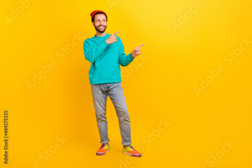 Full size photo of funky guy wear teal sweatshirt red hat directing look at proposition empty space isolated on yellow color background © deagreez