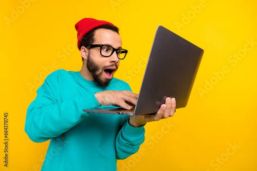 Photo of impressed programmer guy wear teal sweatshirt red beanie hat staring at laptop write code isolated on yellow color background