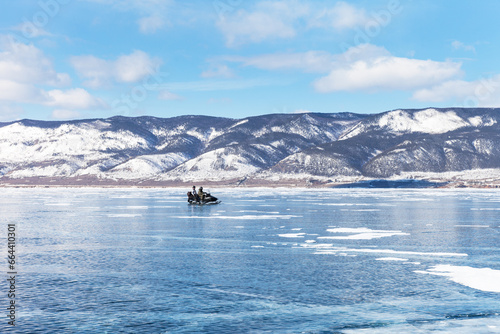 Sunny winter day on frozen Baikal Lake. A couple of tourists travel on the ice of the Small Sea on a snowmobile to Olkhon Island. Winter travel and outdoor recreation