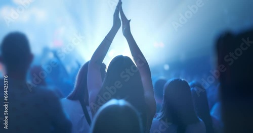 Woman clapping hands at concert, back view. Fan silhouette raises hands up in air, clapping in blue stage spotlights light. Audience applause at music band performance. Happy young girl applauding photo