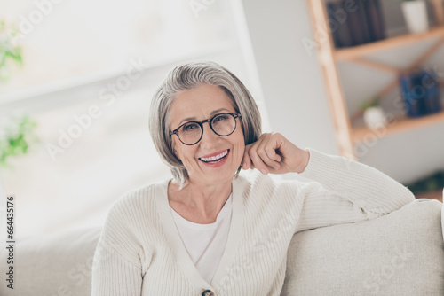 Photo of cheerful good mood elderly lady wear white cardigan spectacles relaxing couch smiling indoors apartment room