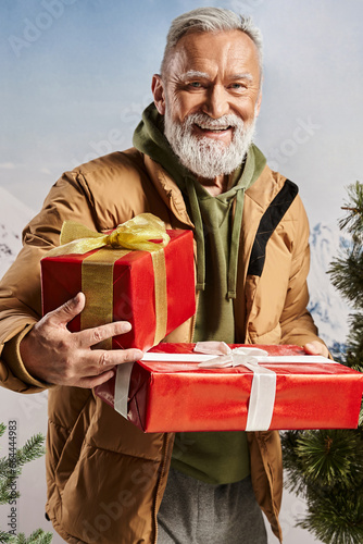 cheerful man dressed as Santa holding red presents and smiling sincerely at camera, winter concept