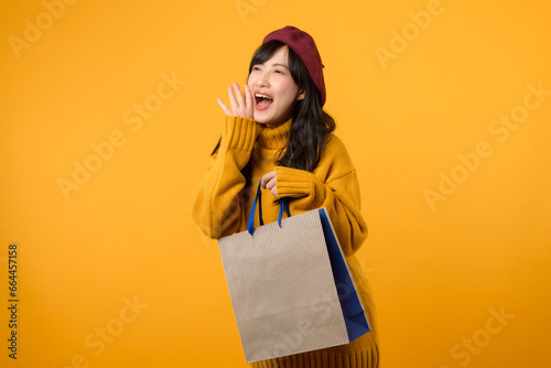 Young Asian woman in her 30s, wearing a yellow sweater and red beret, shouting to free space with shopping paper bag against a vibrant yellow backdrop. photo