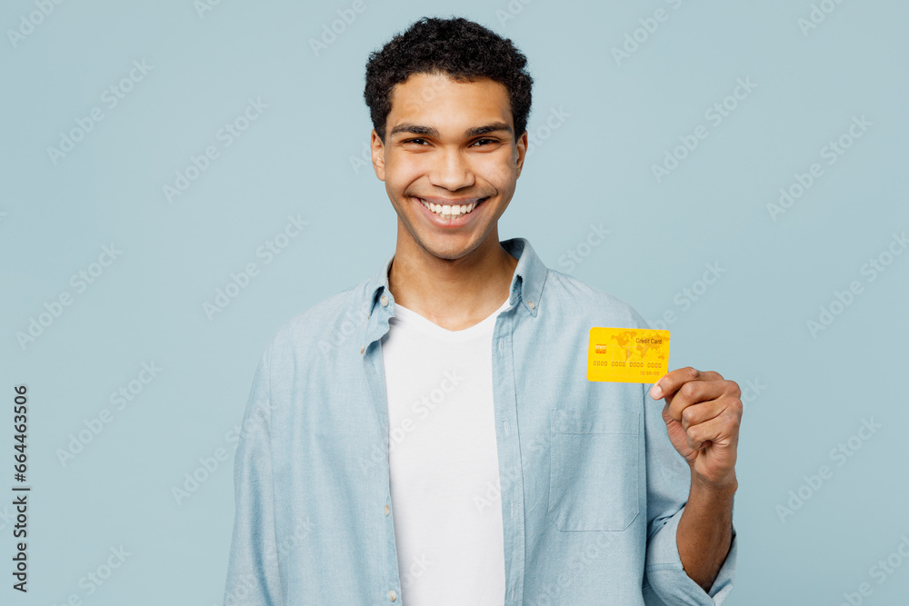 Young cheerful fun smiling man of African American ethnicity he wear shirt casual clothes hold in hand mock up of credit bank card isolated on plain pastel light blue cyan background studio portrait. Young cheerful fun smiling man of African American ethnicity he wear shirt casual clothes hold in hand mock up of credit bank card isolated on plain pastel light blue cyan background studio portrait.