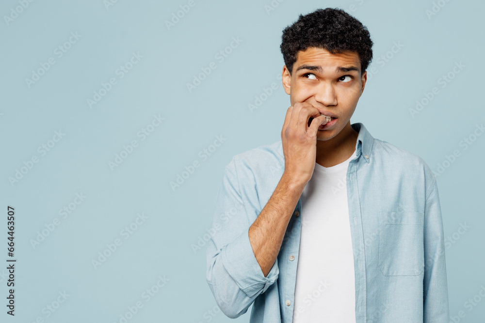 Young sad minded confused man of African American ethnicity wear shirt white t-shirt casual clothes look aside biting nails fingers isolated on plain pastel light blue cyan background studio portrait. Young sad minded confused man of African American ethnicity wear shirt white t-shirt casual clothes look aside biting nails fingers isolated on plain pastel light blue cyan background studio portrait.